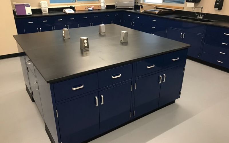 A science lab showcases ADA-compliant lab furniture with a large black-topped island featuring blue cabinets and drawers. Four electrical outlets adorn the island, while the perimeter includes Epoxy resin countertops, sinks, and additional cabinetry for versatile functionality.