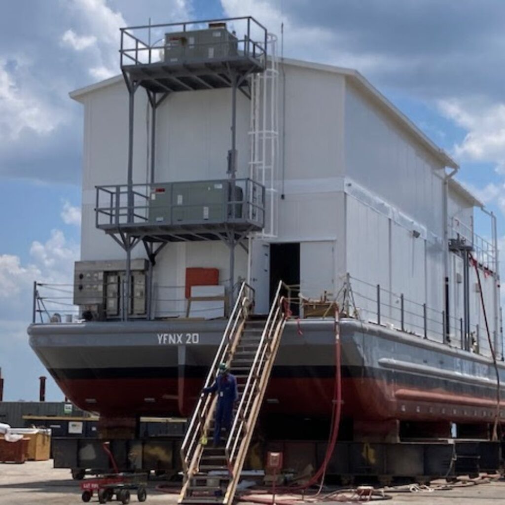 A large modular building resembling a barge-type structure is on land, with multiple stairs leading to the entrance. The vessel, labeled YFNX 20, has a white superstructure and a red hull. Under partly cloudy skies, a person is working near the stairs.