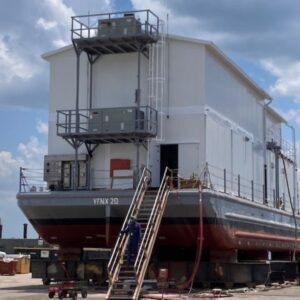 A large modular building resembling a barge-type structure is on land, with multiple stairs leading to the entrance. The vessel, labeled YFNX 20, has a white superstructure and a red hull. Under partly cloudy skies, a person is working near the stairs.
