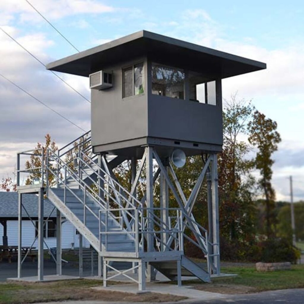 A gray, modular watchtower with a flat roof and windows stands elevated on a metal structure with stairs leading up. It rises against a backdrop of trees and a partly cloudy sky, with a small building visible in the background.