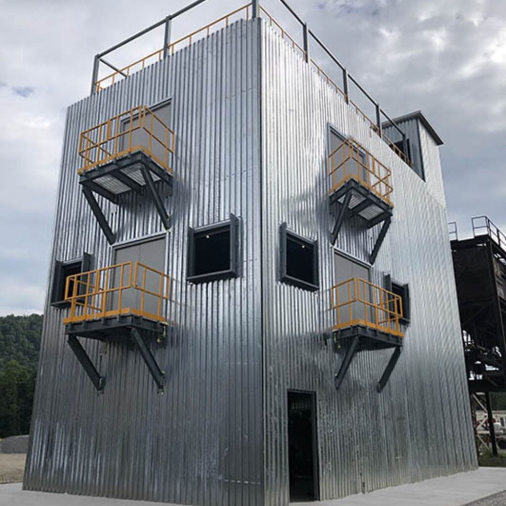 A tall, silver-colored corrugated metal building with multiple orange safety railings on exterior windows, this modern industrial structure resembles modular training towers. It stands against a backdrop of a cloudy sky and distant trees.