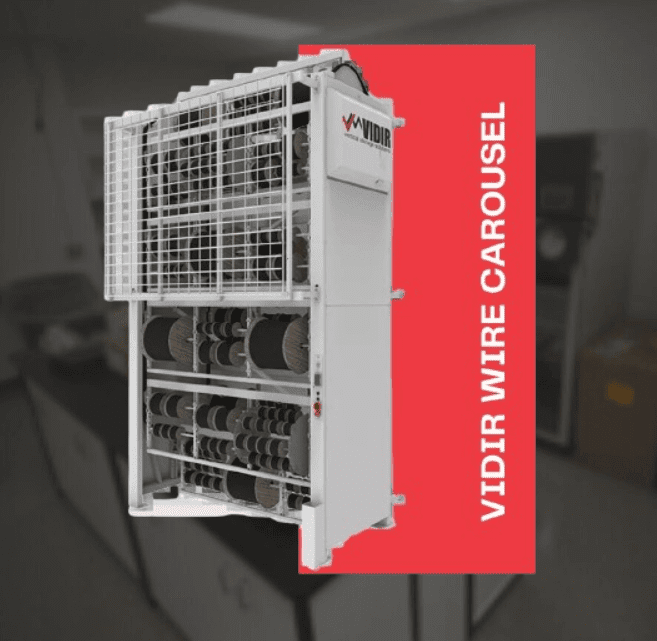 A Vidir Motorized Wire Cable Reel Carousel stands against a blurred indoor background, featuring a red accent panel on the right. The white machine showcases wire spools, with VIDIR WIRE CAROUSEL elegantly displayed in vertical white text.