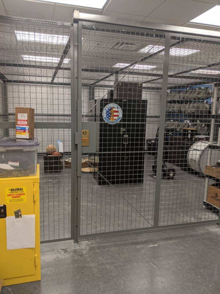 A secure storage area with a wire mesh fence contains shelves and equipment, including a black cabinet with a large emblem on its door. Nearby are cardboard boxes and a yellow storage unit with papers attached. The setting is well-lit.