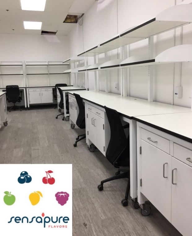 A modern process research lab featuring white workstations, cabinets, and black chairs. The logo in the corner shows colorful fruit icons with the text Sensapure Flavors. The room is well-lit, organized, and exemplifies innovative lab renovation services.