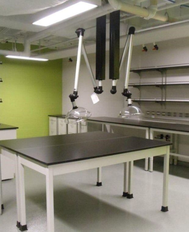 A modern laboratory featuring a green accent wall, equipped with advanced fume extraction systems. Two flexible air extraction arms hang above the black central table. Shelving and cabinets line the walls, ensuring corrosion resistance. The light-colored floor enhances its organized, clean appeal.