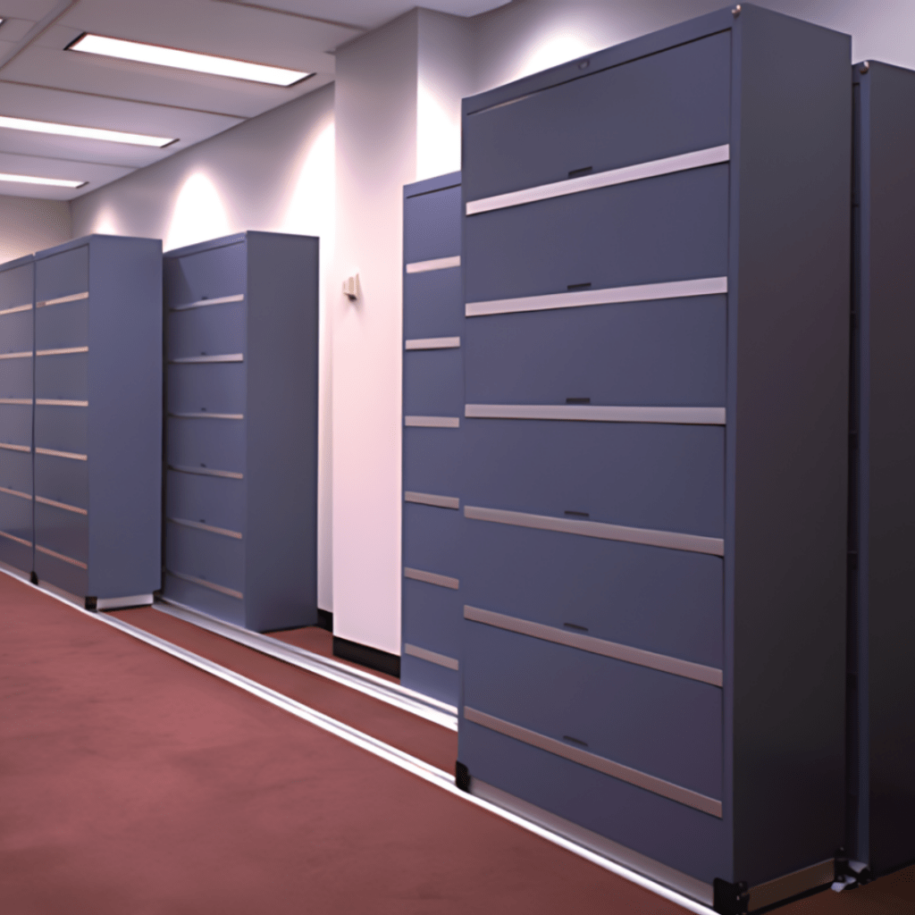A corridor with several closed, gray filing cabinets lines up against cream-colored walls on a red-carpeted floor. The Pipp Mobile Storage Systems cabinets are tall, featuring multiple horizontal drawers, while ceiling lights illuminate the corridor.