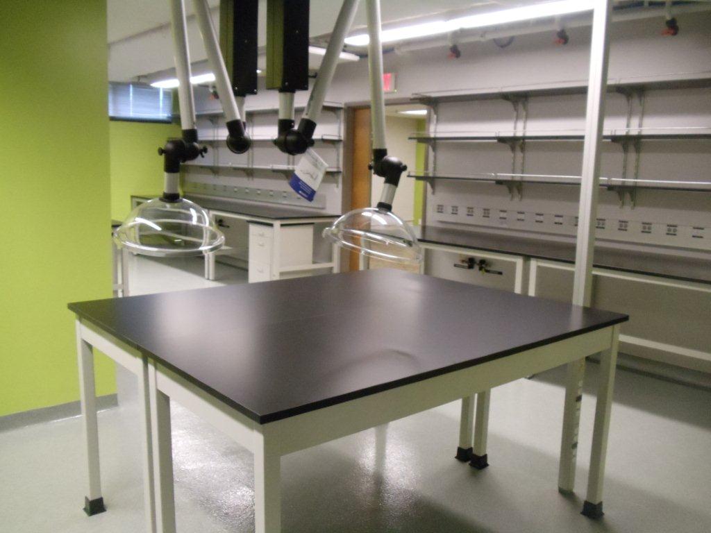 A modern laboratory with lime green walls showcases a central black worktable, overhead ventilation arms, and metal structures. Fume extraction systems ensure adjustable airflow while fluorescent lights illuminate the clean, organized space.