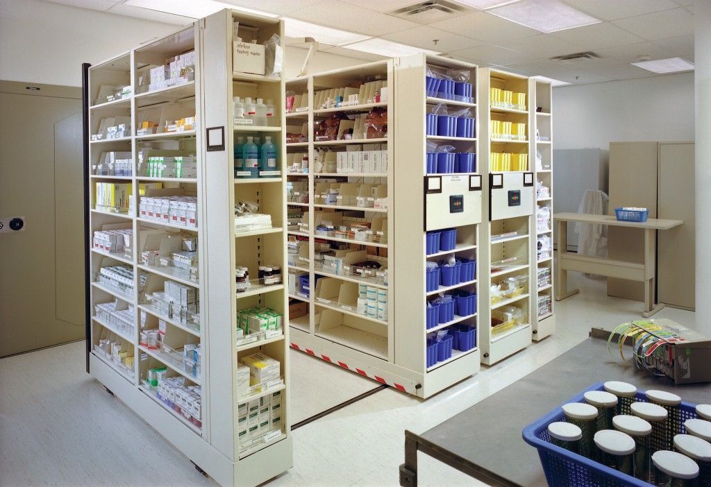 The pharmacy storage room resembles a process research lab, with neatly organized shelves stocked with various medications and supplies. Rows of white shelves hold bottles, boxes, and containers, while a cart with items is visible on the right.