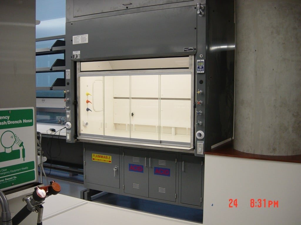 A high-quality lab fume hood with a glass front is shown in a lab setting. It features various controls, warning labels, and storage compartments marked as flammable and corrosive. Nearby, an emergency eyewash station sign underscores the labs enhanced safety measures.
