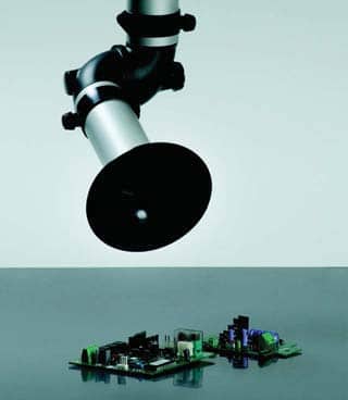 A metallic, angled desk lamp shines light onto various small electronic circuit boards placed on a reflective gray surface, designed with corrosion resistance. The plain light gray background enhances the focus on the lamp and resilient circuit components.