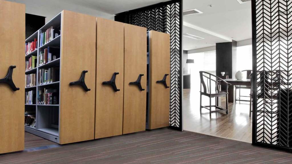 A modern office space with customizable mobile shelving units for lab equipment on the left, featuring wooden panels and black handles. Books are visible on the shelves. On the right, theres a bright area with chairs and tables, separated by decorative black partitions.