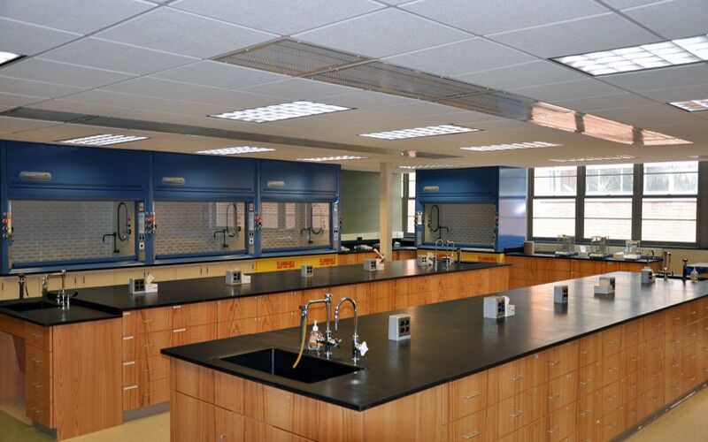 A modern wine chemistry laboratory with specialized lab furniture, featuring long black countertops, wooden cabinets, and multiple workstations. Chemical fume hoods line the walls while large windows let in natural light. The lab layout is well-organized and equipped for experiments.