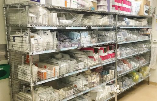 A metal shelf unit, emblematic of a process research lab, holds an array of medical supplies, neatly arranged in rows. Situated against the wall in a clinical setting, it reflects precision and organization amidst an environment dedicated to meticulous scientific exploration.