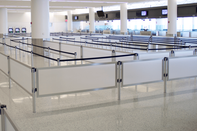 An empty airport check-in area features ADA-compliant queues with multiple row dividers and retractable barriers guiding passengers. Four check-in counters with screens are visible in the background, all under bright lighting and a clean, modern atmosphere.