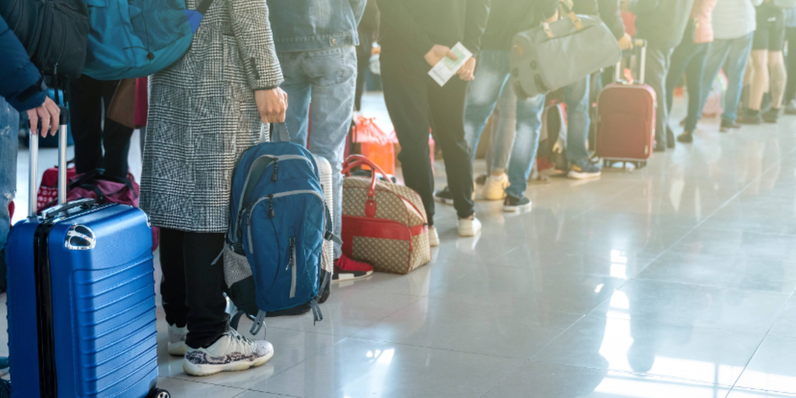 In an ADA-compliant queue at the airport terminal, people stand in line, each clutching their luggage. A blue suitcase stands out among backpacks, while sunlight dances off the polished floor.