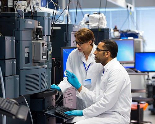 Two scientists in white lab coats and blue gloves work diligently in a laboratory, analyzing data on a computer linked to large equipment. With environmental chamber storage solutions in play, one sits while the other stands amid computer monitors and various lab apparatus in the background.