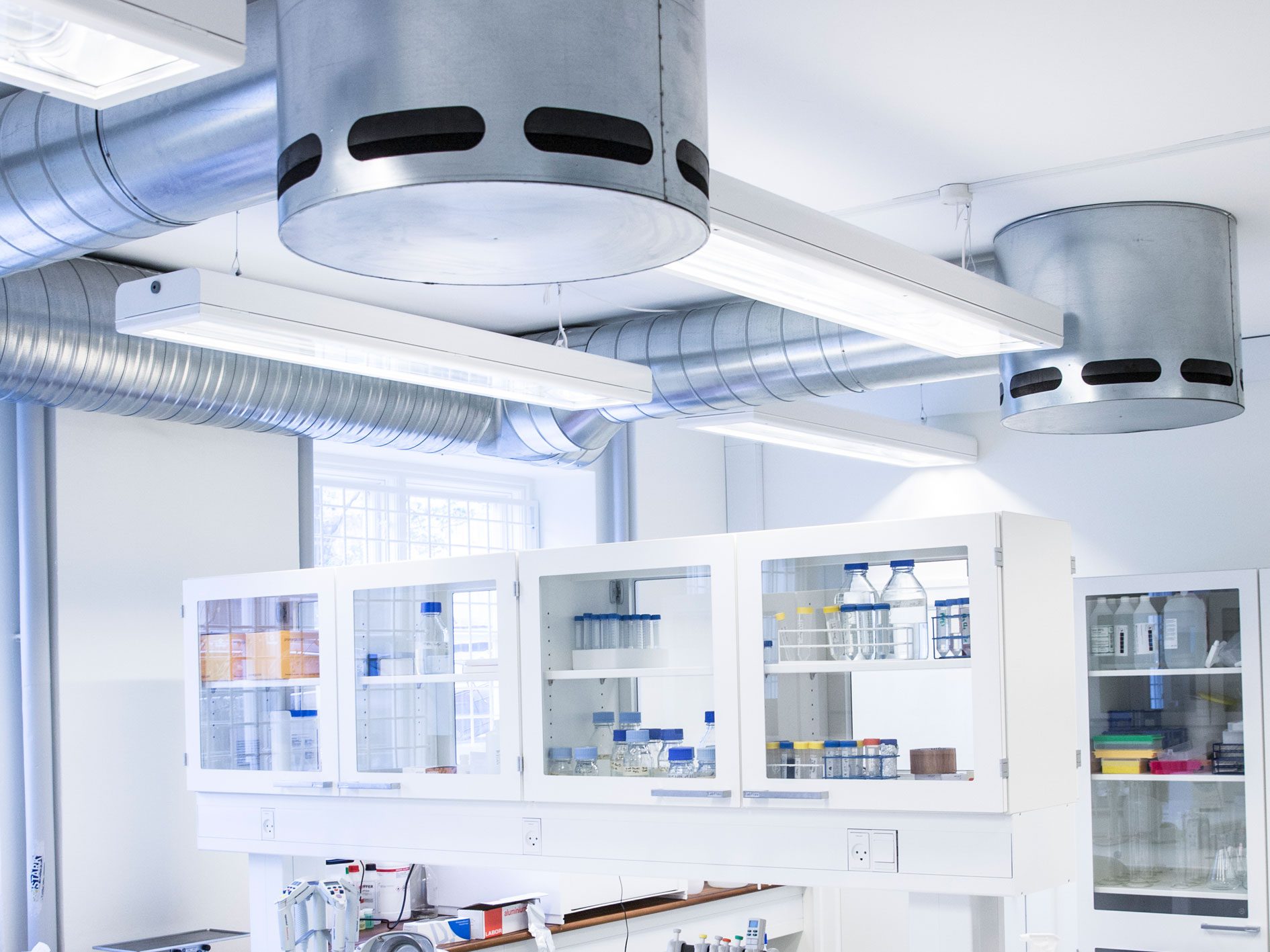 The modern laboratory interior features white cabinets and glass doors housing various bottles. Large metallic ventilation ducts adorn the ceiling, accompanied by fluorescent lights. The room, designed with advanced storage solutions, appears bright and clean, ideal for environmental chamber setups.