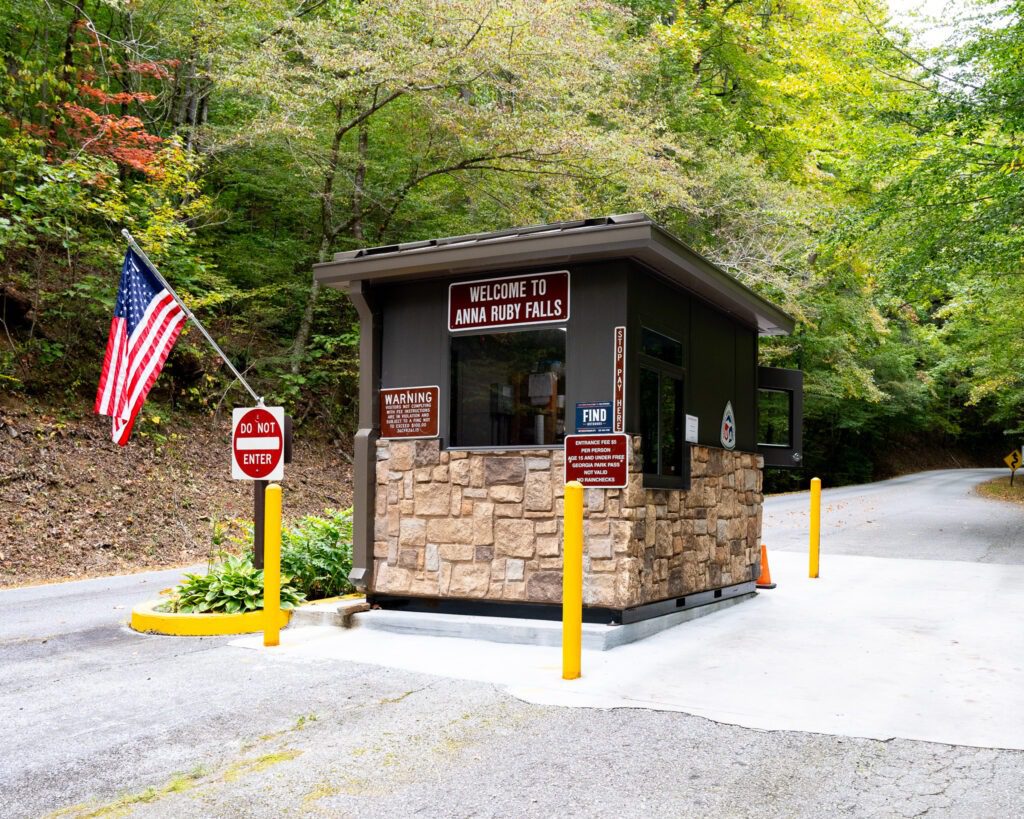 A small entrance booth for Anna Ruby Falls stands by a forest road, reminiscent of classic guard booths in stock. It features a stone base, brown walls, and a sign above. An American flag flutters near a Do Not Enter sign, amid green trees and the curving road.