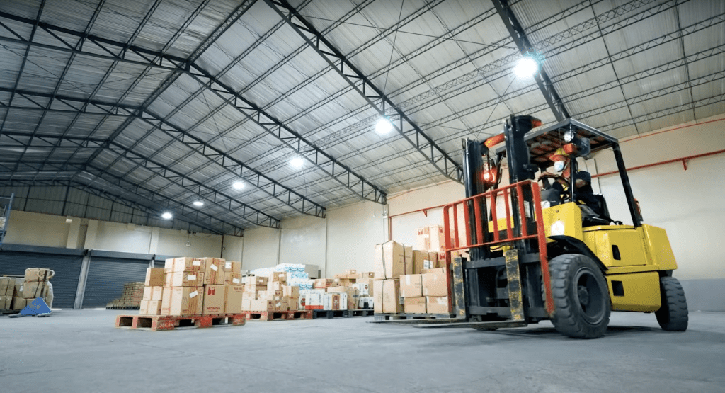 In the well-lit, expansive warehouse with high ceilings, a forklift operator maneuvers a pallet of boxes. More pallets await in the background. The concrete floor and light beige walls showcase efforts to improve warehouse designs for enhanced efficiency and aesthetic appeal.