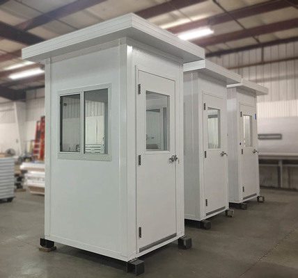 Three small, white security booths with flat roofs and windows on one side are lined up indoors. These elevated guard booths, in stock now, have doors with handles. The background features industrial elements like beams and equipment, complementing their sturdy metal legs.