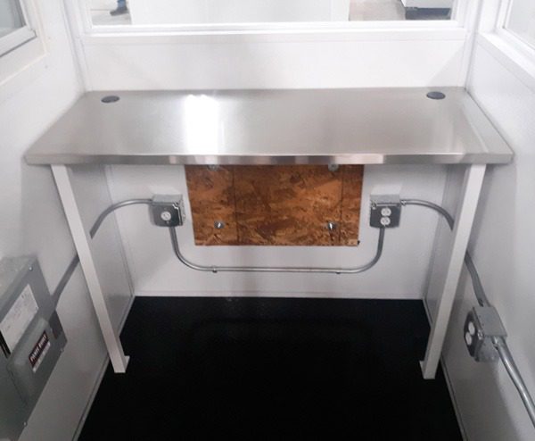 In this compact space, a stainless steel desk contrasts the black floor. Equipped with electrical outlets and exposed conduit piping, it mirrors the practicality of guard booths in stock. The rooms white walls and a large window brighten the utilitarian setup.