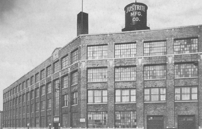 Historic brick industrial building with large square windows, featuring a rooftop water tower boldly showcasing the Justrite MFG. CO. The architecture beautifully reflects early 20th-century factory design, set against a backdrop of a cloudy sky.