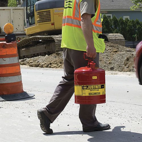 A construction worker in a high-visibility vest walks along a paved road, carrying a red Justrite safety container. An excavator and orange traffic barrels stand prominently in the background, illustrating the busy work site.