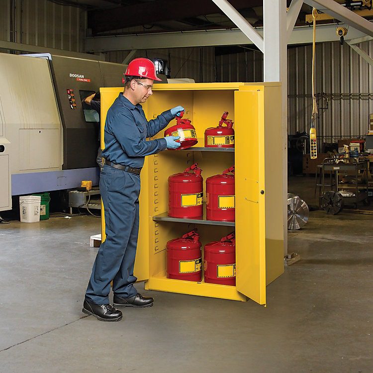 A worker, equipped with a red hard hat and blue work uniform, places a red safety can into a Justrite Safe Chemical Storage cabinet. The open yellow cabinet reveals several more red safety cans inside, set against a backdrop of industrial machinery and equipment.