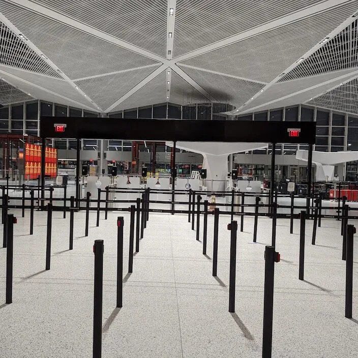 The empty airport security checkpoint features Visiontrons expertly arranged black stanchions in a zigzag pattern, promising a seamless, efficient queue experience. Modern architectural ceiling design dazzles with triangular shapes, while bright overhead lights and red exit signs complete the scene.