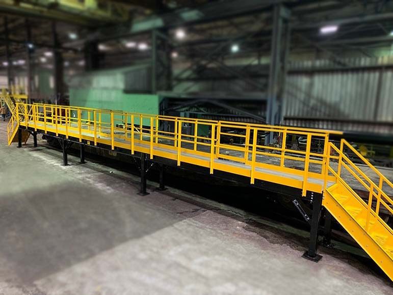 A long industrial yellow catwalk with railings, supported by black metal legs, stretches across a factory floor. A staircase leads up to one end of the platform. The background features blurred industrial equipment and structures.