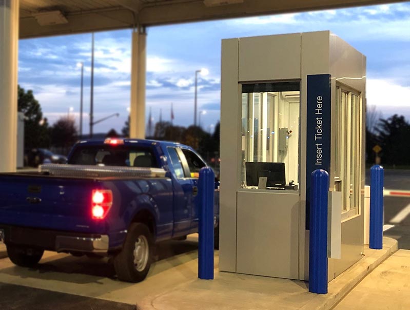 A blue pickup truck is halted at a parking entrance guard booth in stock as dusk settles. The booth, surrounded by blue bollards for safety, displays a sign reading Insert Ticket Here. Trees and a partly cloudy sky create a serene backdrop.