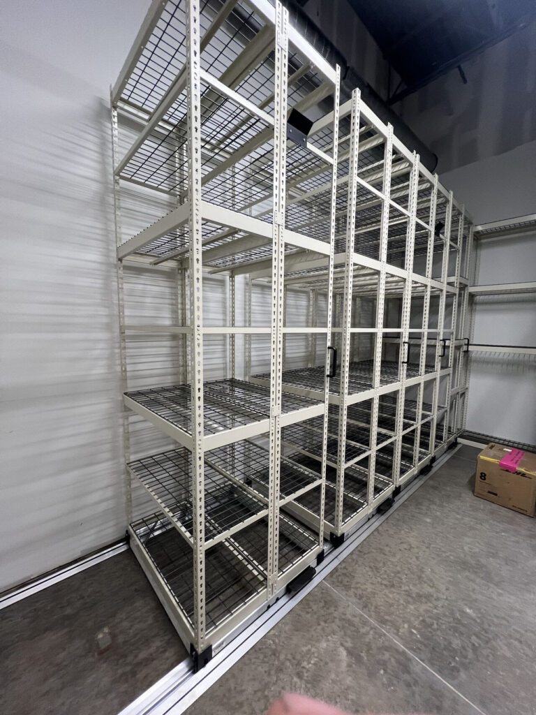 A large, empty metal shelving unit stands ready for shoe inventory management in a warehouse setting. The shelves feature a wire grid design, complementing the industrial space with its concrete floors and high ceiling. A lone cardboard box is visible on the right side.
