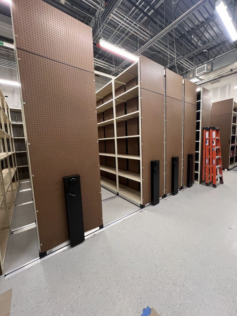 Empty movable shelving units on tracks in a warehouse or storage room, ready for efficient shoe inventory management. The shelves have brown pegboard sides. A red ladder is visible nearby, with an overhead view of the ceilings exposed pipes and lights.