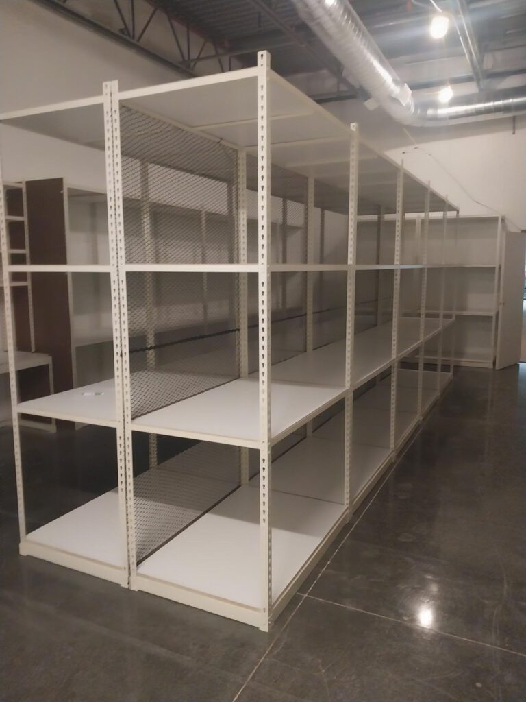 Empty white metal shelving units in a warehouse setting, designed for efficient shoe inventory management, stand on a polished concrete floor. Overhead, industrial lighting and an exposed ceiling with visible ductwork can be seen. A mesh panel is attached to one of the shelves.