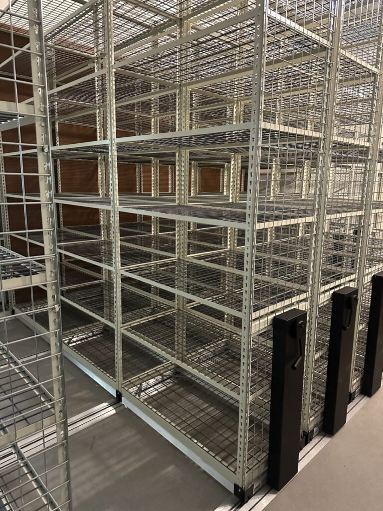Metallic storage shelves with a mesh design are in a spacious room, ideal for shoe inventory management. Mounted on tracks for mobility, they feature black rotating handles to adjust positions. Wooden walls and concrete flooring enhance the industrial look.