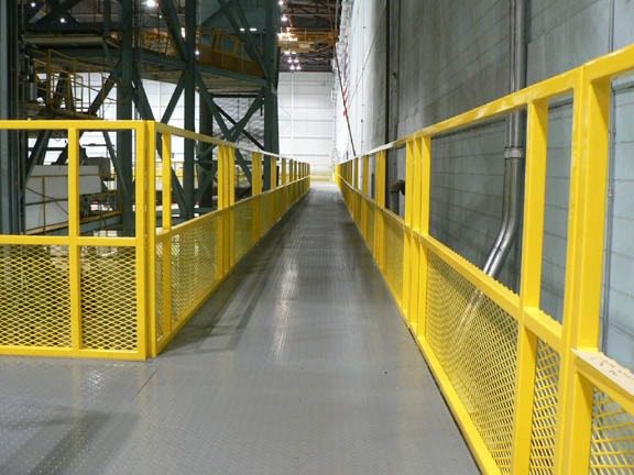 A long industrial catwalk with yellow metal railings on both sides stretches out. The metallic gray floor leads to a spacious, well-lit interior with high ceilings and visible structural beams.