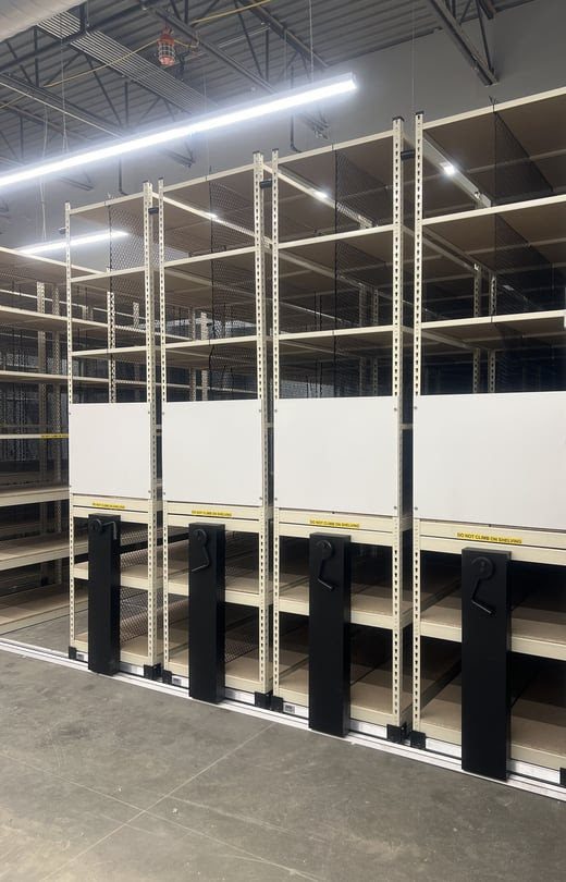 A photo of a stockroom solutions setup showcases large, empty mobile shelving units. These shelves, with handles for lateral movement, efficiently save space. The high-ceilinged room glows under overhead fluorescent lights, while the concrete floor completes the practical look.