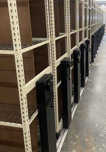 A row of mobile shelving units with hand cranks stands ready in the warehouse, exemplifying innovative stockroom solutions. The empty shelves are perfectly aligned on floor tracks, designed to slide and save space. Overhead, industrial lighting highlights the concrete floor below.
