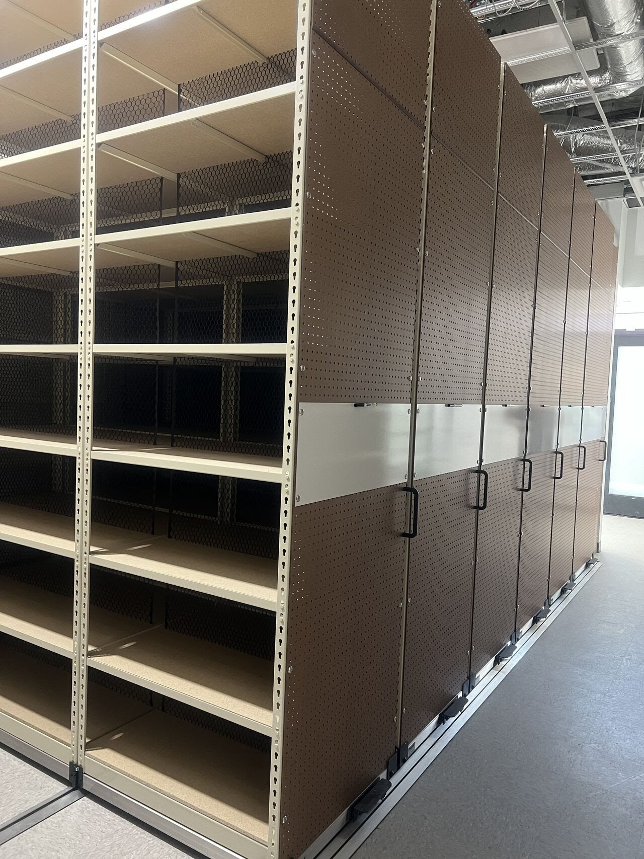 A large, empty shelving unit with multiple shelves stands in a room. This Stockroom Solutions piece is crafted from wood panels in a metal frame and features black mesh sides. The unit is on wheels, allowing it to be moved along tracks on the floor effortlessly.