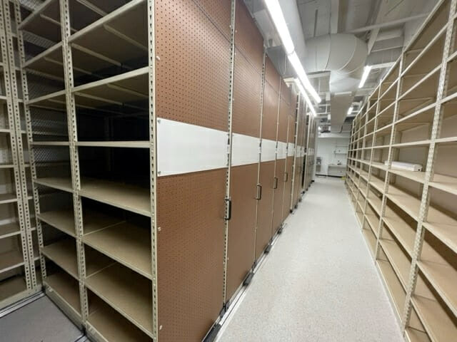 A wide-angle glimpse into a pristine stockroom solutions area reveals rows of beige shelves on both sides. These shelves, metal-framed with wooden surfaces, flank a brown partition in the middle. The gray floor and ceiling with exposed pipes complete this organized space.