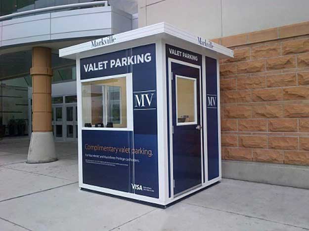 A small rectangular valet parking booth with dark blue panels and white text is stationed outside a building, featuring Valet Parking and Complimentary valet parking. Similar to our guard booths in stock, it comes equipped with windows and an entrance door.