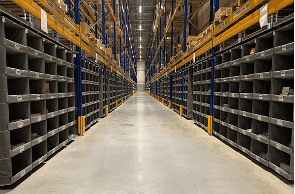 A warehouse aisle with rows of gray storage bins on either side features optimized towering shelves filled with brown boxes. The concrete floor is clear, and the overhead lights brightly illuminate the well-organized space.