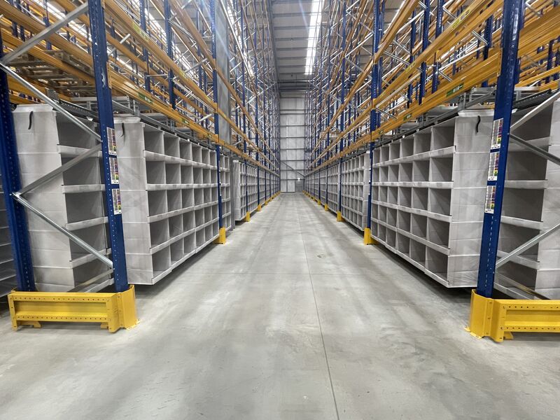 An empty warehouse aisle with tall yellow and blue metal shelving units on either side showcases the potential for efficient warehouse optimization. The clean concrete floor and evenly spaced ceiling lights illuminate the spacious area, ready to boost productivity.