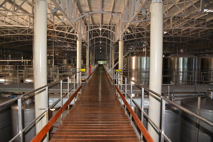 A long, narrow wooden walkway with metal railings runs through an industrial facility, resembling a custom catwalk system. Large stainless steel tanks and pipes are visible on either side, with the ceiling supported by a network of robust metal beams.