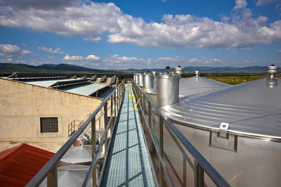 From the rooftop walkway, equipped with a custom catwalk system, overlook expansive metal tanks set against scenic hills and a partly cloudy sky. Safety railings line both sides of this industrial vantage point.