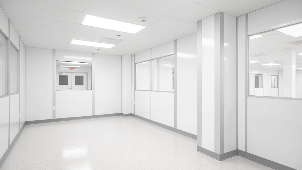 An empty, pristine hallway in a sterile, white laboratory or cleanroom. The walls and ceiling gleam under bright fluorescent lighting. Large windows line the corridor, allowing visibility into adjacent rooms where Gowning Room Essentials are meticulously stored and organized.