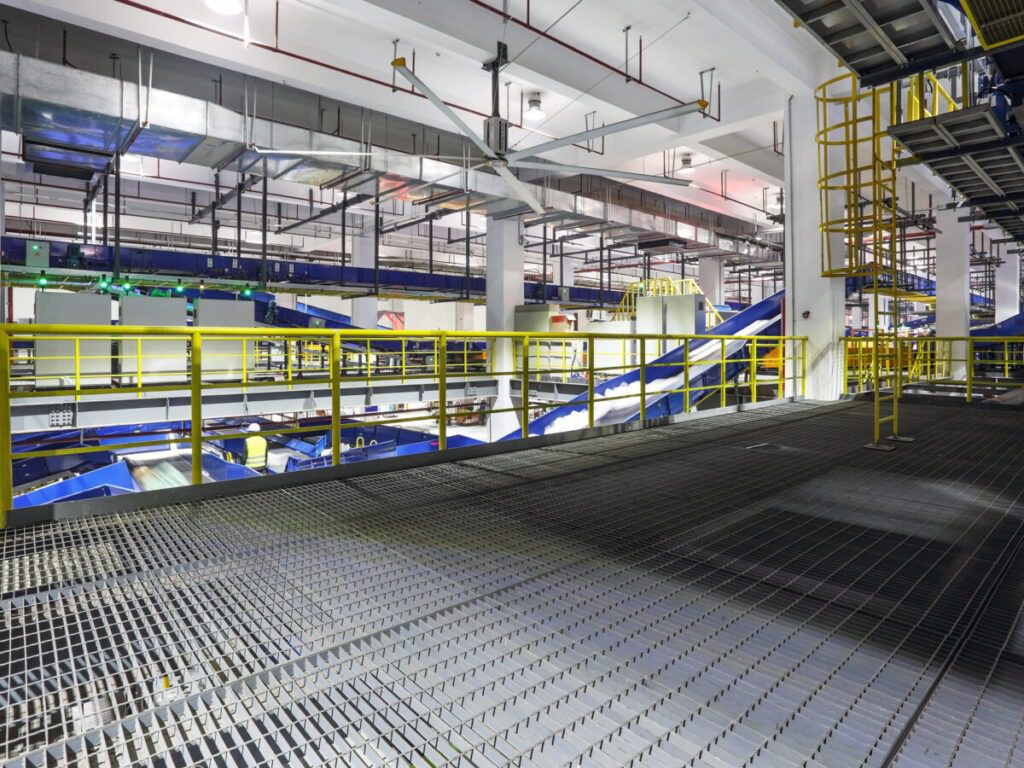The interior of the large industrial facility features metal walkways with yellow safety railings. Overhead piping and conveyor systems snake above a custom catwalk system, ensuring seamless movement throughout the well-lit, spacious, and organized environment.