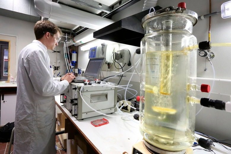 A person in a lab coat expertly navigates their laptop amidst scientific equipment and a large glass container with liquid and tubing, ensuring compliance and safety in the laboratory. The room is meticulously designed, filled with cables and instruments for optimal functionality.