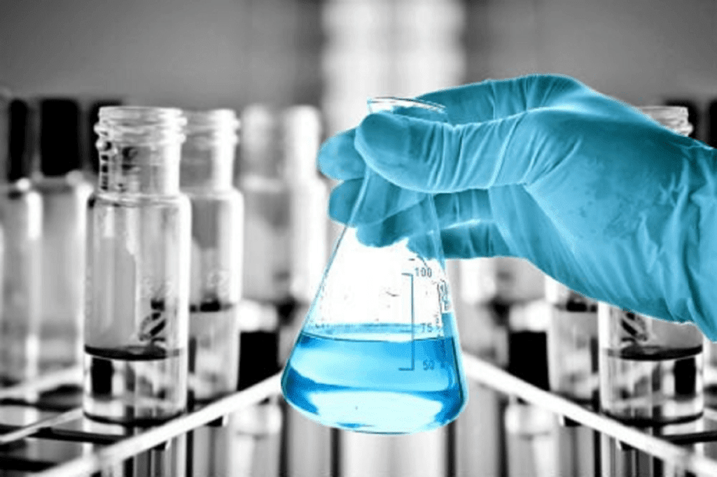 A gloved hand holds a conical flask filled with blue liquid in a laboratory setting, underscoring expert laboratory compliance and safety consulting. The blurred background features test tubes in a rack, highlighting the flask as the images focal point.