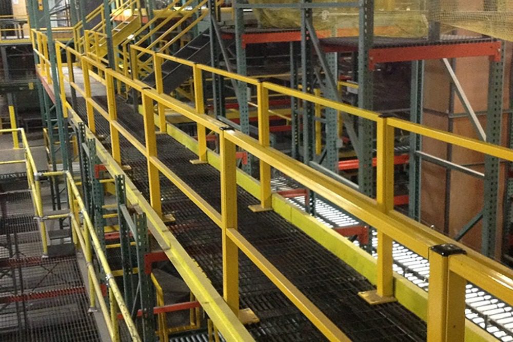 Yellow metal walkways and railings form a custom catwalk system in an industrial warehouse, with shelving units neatly arranged. Stairs leading to another level can be seen in the background, enhancing the streamlined functionality of the space.